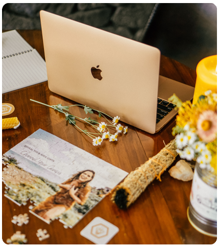 A laptop sits on a wood table, surrounded by flowers, a candle, and a bundle of sage