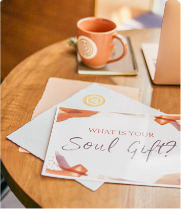 an orange coffee mug sits on a wooden table with a postcard that reads "What is your soul gift"