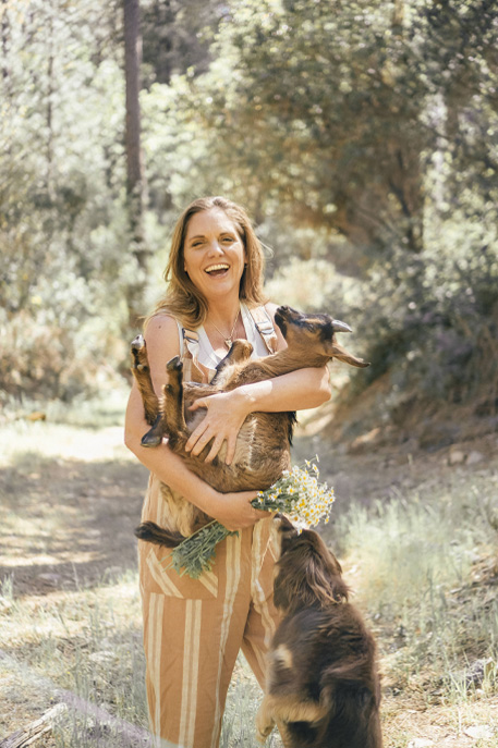 Mellissa is standing in the woods with a smile on her face and holding a baby goat in her arms