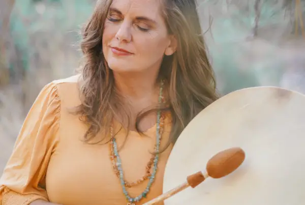 A woman stands outdoors holding a large drum and drumstick while her eyes are closed. She is wearing a yellow top and a beaded necklace, with a pine tree visible in the background.