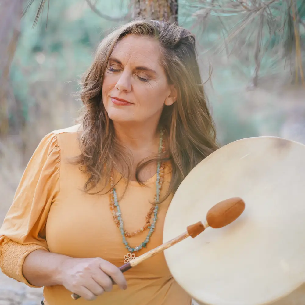 A woman stands outdoors holding a large drum and drumstick while her eyes are closed. She is wearing a yellow top and a beaded necklace, with a pine tree visible in the background.