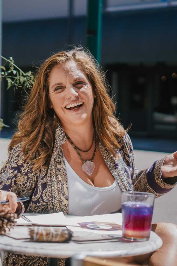 A woman sitting outdoors at a small table while viewing the wheel of wisdom. She has an ornate coverup over a white tank top and a beaded necklace on. In front of her is a fancy purple, red and orange drink and the sun is shining in the background.