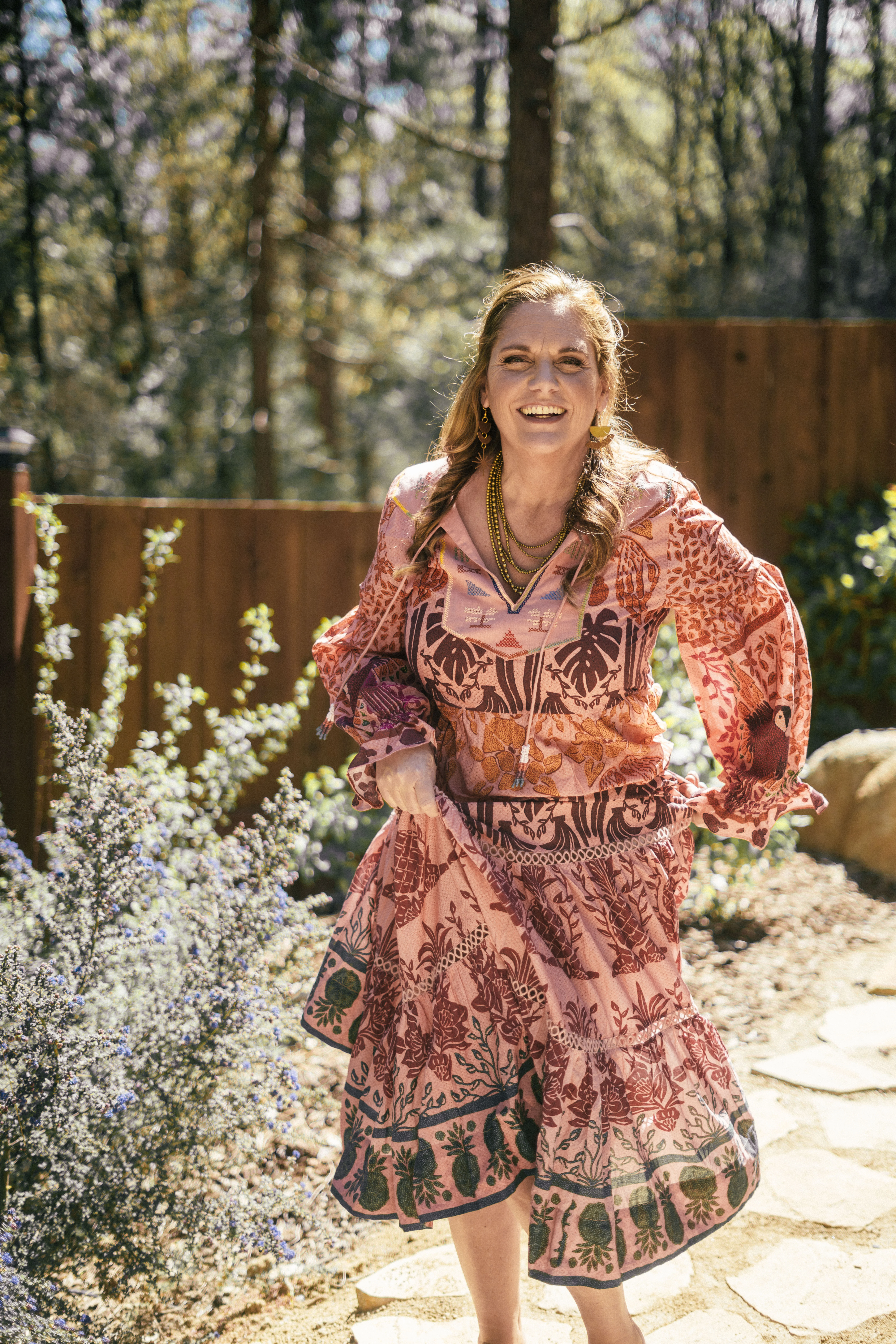 A woman with long brown hair wearing a bright red ornate dress stands on a stone path surrounded by trees, posing with her in a relaxed lean as she is lifting her dress and smiling in the sunshine. There are lots of trees and foliage surrounding her in the background.